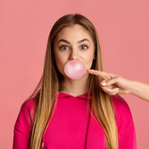 a woman wearing a pink hoodie blowing a bubble with a hand pointed to pop the bubble