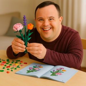 A man with down syndrome sitting at a table holding a floral lego project with the instructions open on the table in front of him