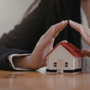 a mini house with a person's hands above it protecting it