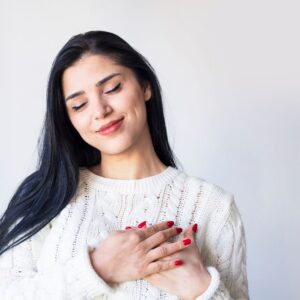 Person with long dark hair wearing a light knit sweater, eyes closed and hands over the chest with red-painted fingernails, suggesting calm