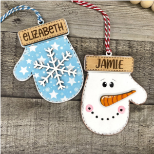 Two painted holiday mittens hanging on a wooden surface: a blue mitten labeled “ELIZABETH” and a white mitten-shaped figure labeled “JAMIE” with a carrot nose and red-and-white string.