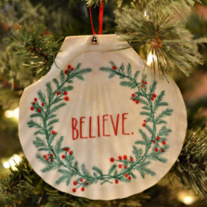 A sea shell ornament with Christmas greenery and small lights, featuring the word "believe" in red lettering.