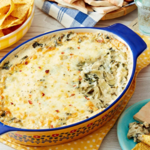 Blue-edged ceramic baking dish filled with a baked spinach, artichoke and herb casserole, topped with melted cheese and browned spots, set on a wooden table with tortillas, a striped cloth, and a small plate nearby.