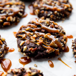 Close-up of several chocolate cookies topped with chopped nuts and a drizzle of caramel, on a light surface.