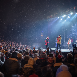 Crowd packed into a concert venue as three performers sing and dance on a brightly lit stage, surrounded by blue spotlights, haze, and falling confetti.