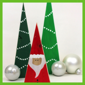 A white square centered on a solid green background, with 2 wooden triangles painted like Christmas trees with white trim and a smaller triangle painted red and white to look like Santa with silver Christmas ornaments surrounding them.
