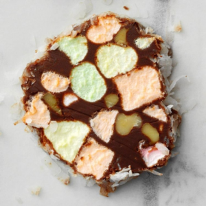 A round chocolate cookie slice with visible pale yellow and light green marshmallow pieces, scattered coconut around the edges, on a light gray surface.