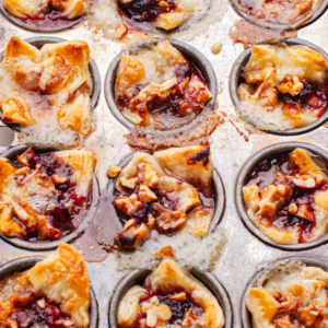 Mini baked tartlets in round tins, each topped with golden pastry, dark fruit filling, and scattered crumbs.
