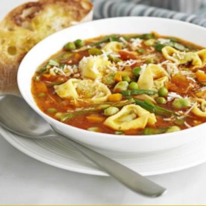 A white bowl of hearty pasta soup with tomatoes, green peas, sliced vegetables, and shredded cheese, served with a spoon and a round bread roll on a striped cloth.
