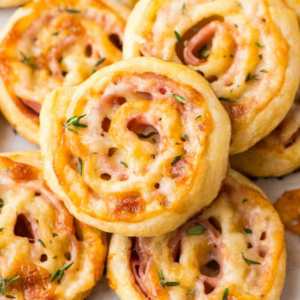 Golden mini pinwheels stacked together, each filled with melted cheese, visible ham slices, and tiny green herb flakes.