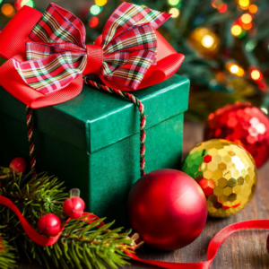 Green gift boxes tied with red plaid bows, surrounded by red ornaments, pine branches, and a shiny gold bauble on a wooden surface with colorful holiday lights in the background.