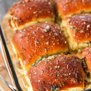 Close-up of golden-brown bread rolls in a glass baking tray, topped with flaky salt and herbs, with one roll in the foreground showing a browned, textured crust.
