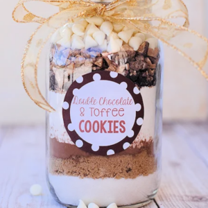 A glass jar filled with cookie mix layers and chocolate chunks, tied with gold ribbon; a label reads “Cookie Chocolate & Toffee COOKIES.”