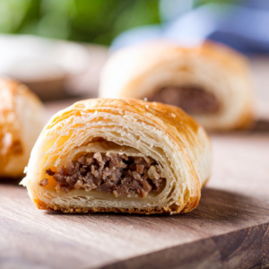 A close-up of a sliced pastry with flaky layers and a dark ground pork filling, resting on a wooden surface, with more pastries blurred in the background.