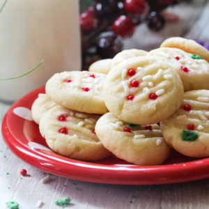 A red plate piled with butter cookies topped with white icing and red sprinkles, with a few crumbs on a light tabletop and blurred holiday decorations in the background.