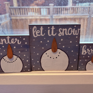 Three wood blocks with snow man faces sitting a window sill with text on them that is winter themed "winter" "let it snow" and "brr."