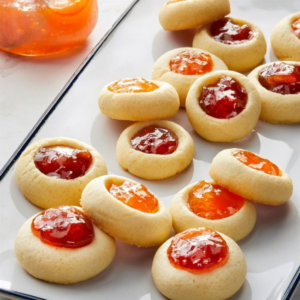 Tray of small butter cookies with thumbprint wells filled with red and orange jam, with a jar of jam in the top left corner.
